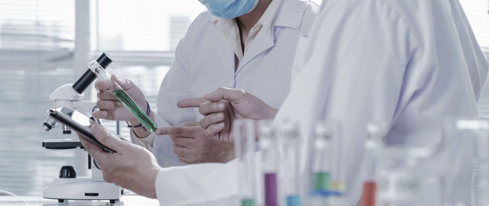 Close-up shot of unrecognizable chemists wearing white coats examining content of test tube and taking necessary notes with help of digital tablet while carrying out experiment